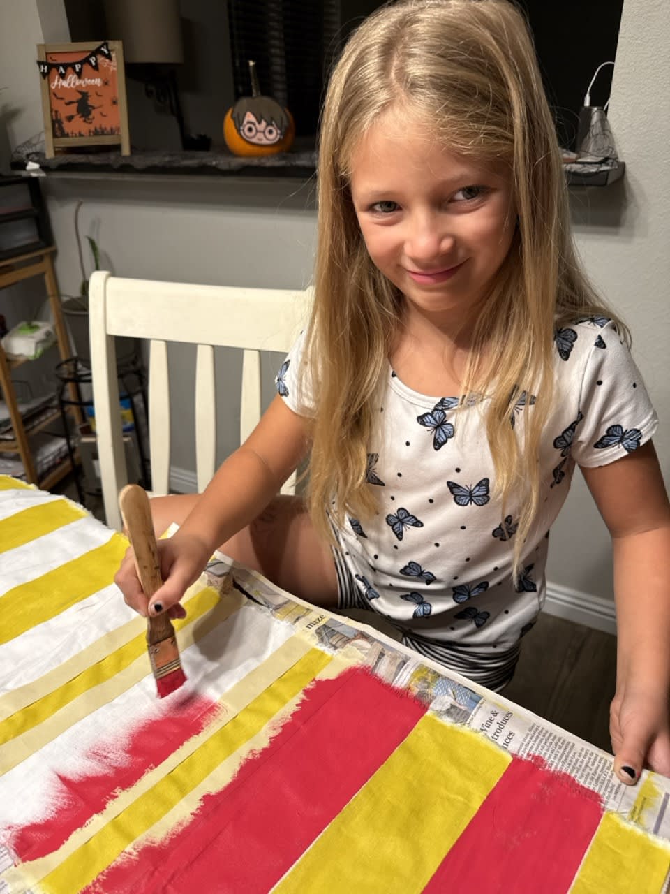 With a mischievous grin, a child paints a component for her own Halloween costume at a cluttered home dining room table