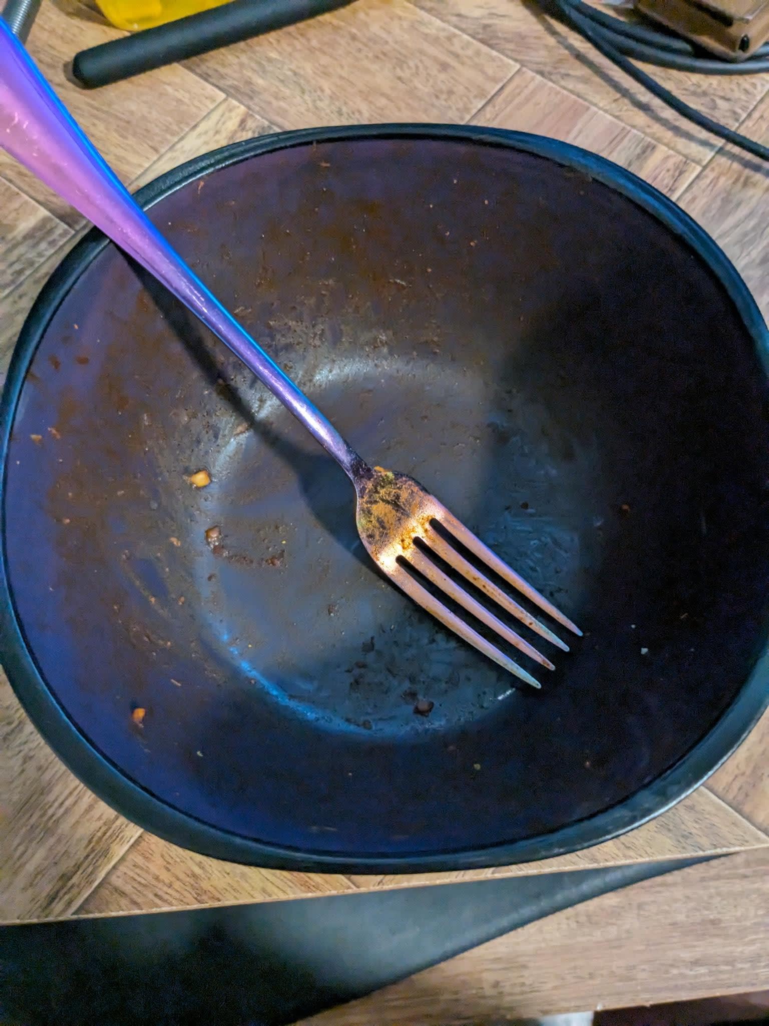 An overhead view of a blue bowl shaped like a square with rounded edges and corners. The bowl is empty except for a fork. Both the fork and bowl are covered in a residue of dried, orange Buldak sauce.