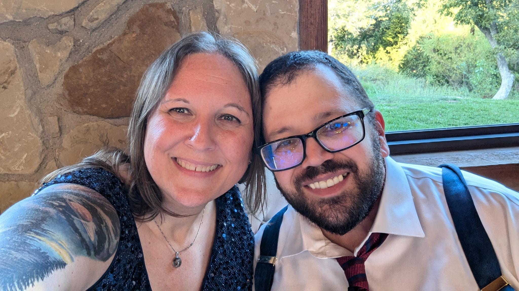 A woman with brown shoulder-length hair, hazel eyes, and a sleeve tattoo on her selfie arm. She is dressed in a fancy blue sleeveless dress and leaning into a man with short brown hair, full beard, and mustache wearing chunky black glasses and suspenders over a white dress shirt with a loosened tie. Both are seated and leaning into each other with big goofy smiles and looking at the camera.