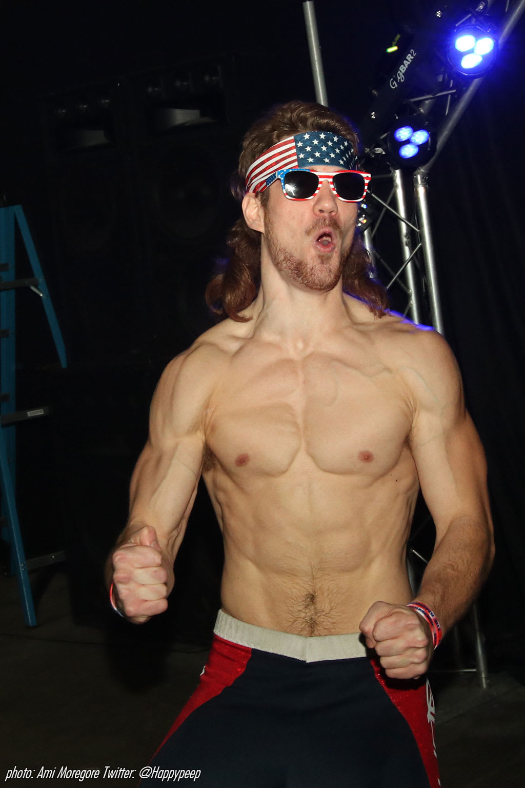 Mike Quay makes his entrance - a white man with brownish red hair, an american flag headband, sunglasses and no shirt showing his defined physique