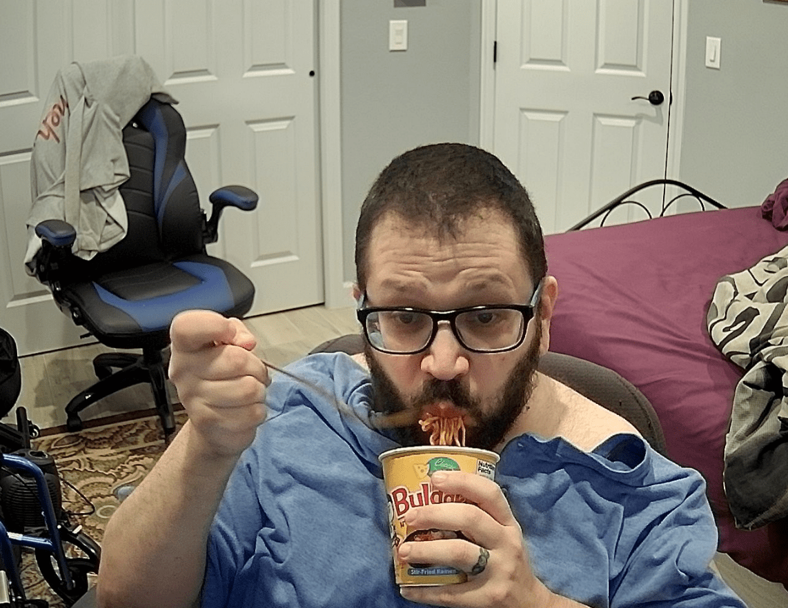 A bearded man in his 40s with brown hair and wearing black, thick-rimmed glasses. He is shoveling a forkful of noodles in his mouth from a Samyang cup he's holding under his mouth. A blue shirt is obviously draped on top of his chest and arms instead of being worn as normal. A gray hoodie with "meh!" on the back is carelessly slung over the office chair in the background.